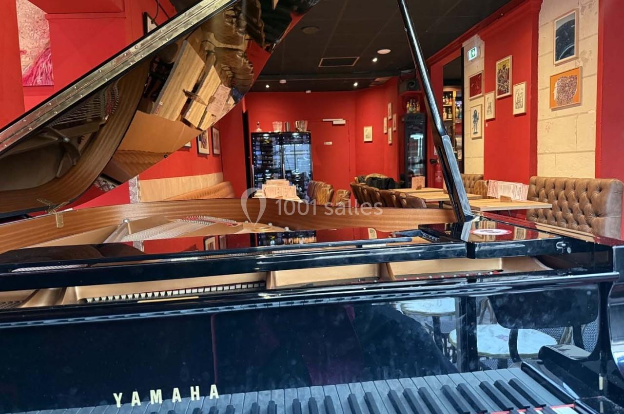 Piano à queue noir dans un restaurant aux murs rouges, avec des tables dressées et des fauteuils en arrière-plan.
