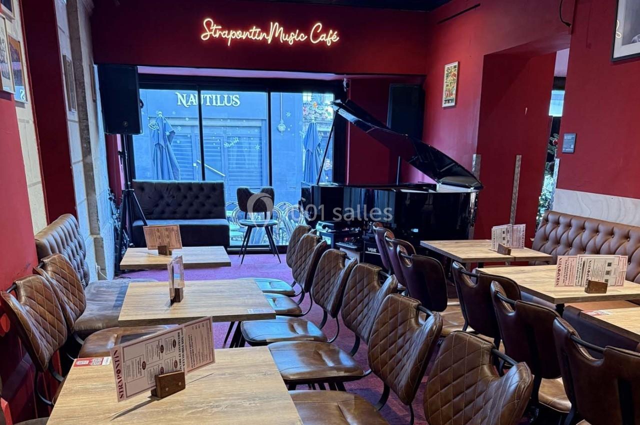 Salle de café avec des tables en bois, des chaises rembourrées et un piano à queue devant une baie vitrée.
