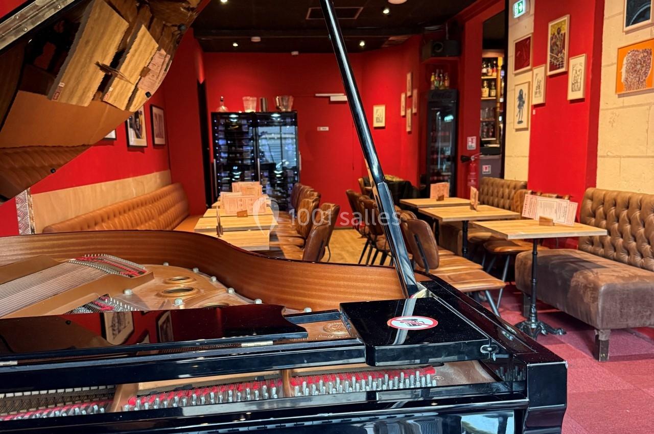 Salle de restaurant avec murs rouges, tables alignées, banquettes en cuir et un piano à queue au premier plan.