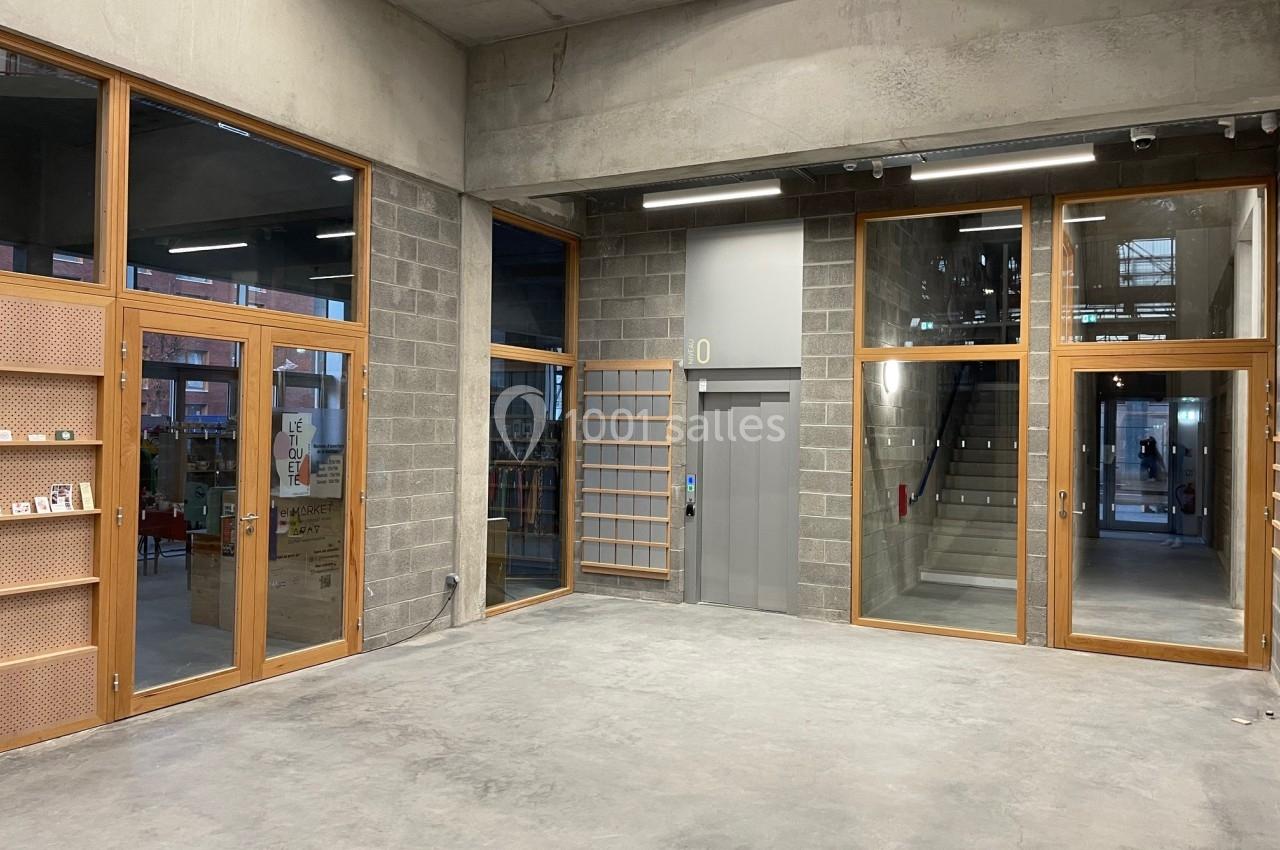 Intérieur d'un bâtiment moderne avec murs en béton, grandes baies vitrées, un ascenseur et un escalier visible au fond.