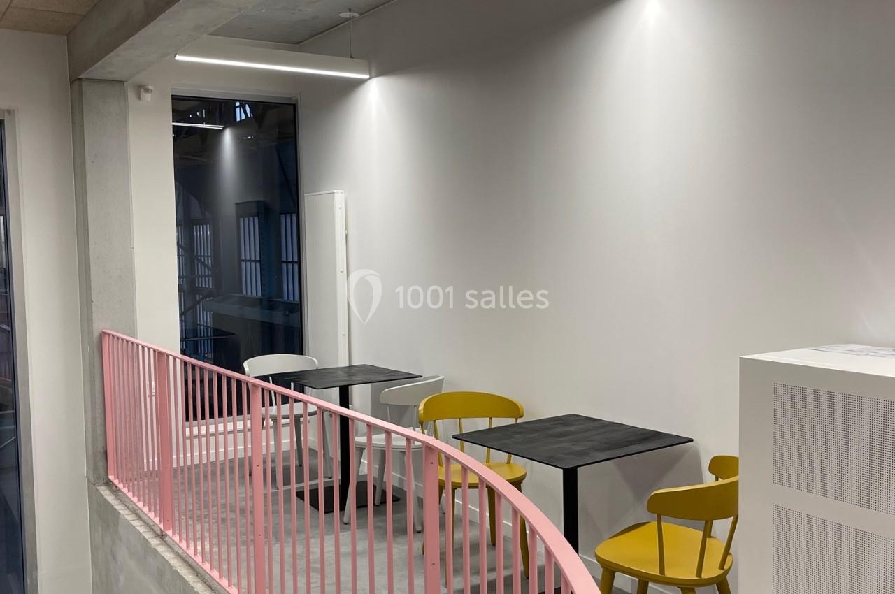 Espace mezzanine avec tables noires, chaises jaunes et rambarde rose, éclairé par des lampes suspendues.