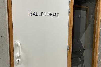 Salle de classe moderne avec tables en U, chaises en bois, sol en béton et éclairage suspendu.