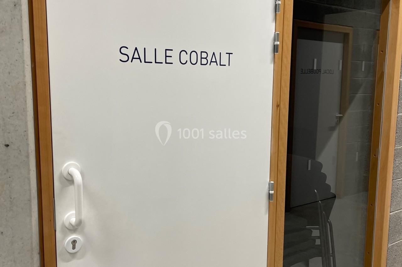 Porte blanche avec inscription ’Salle Cobalt’, encadrée de bois, à côté d'une paroi vitrée donnant sur une salle.