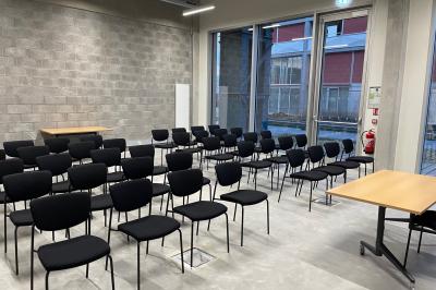 Salle de classe moderne avec tables en U, chaises en bois, sol en béton et éclairage suspendu.