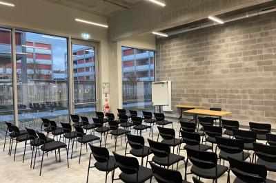Salle de classe moderne avec tables en U, chaises en bois, sol en béton et éclairage suspendu.