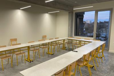 Salle de classe moderne avec tables en U, chaises en bois, sol en béton et éclairage suspendu.