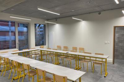 Salle de classe moderne avec tables en U, chaises en bois, sol en béton et éclairage suspendu.