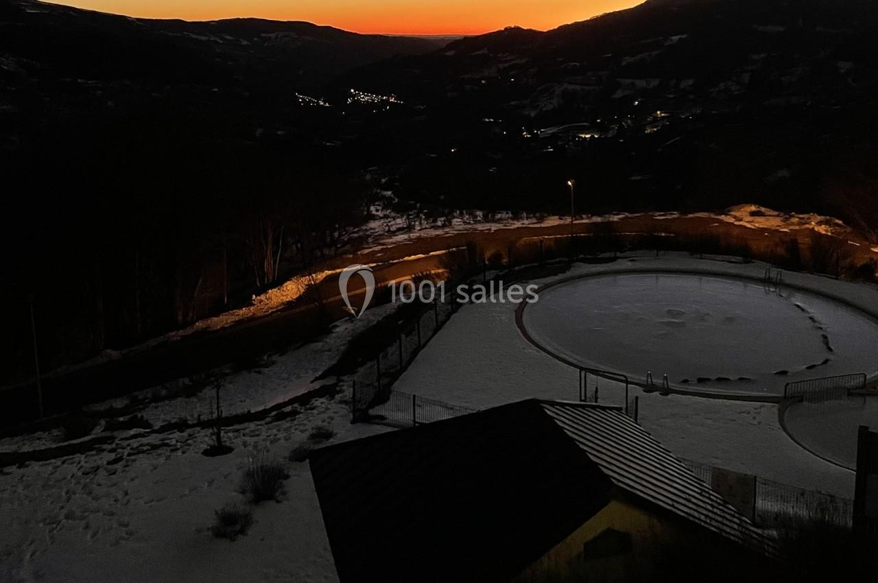 Vue nocturne d'un paysage enneigé avec un ciel orangé, des collines et des lumières urbaines au loin.