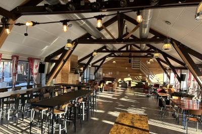 Salle de restaurant lumineuse avec poutres apparentes, grandes fenêtres, tables en bois et chaises hautes.