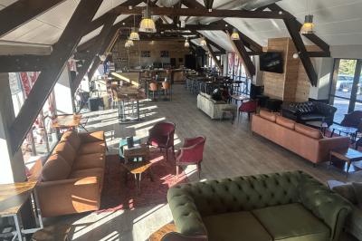 Salle de restaurant lumineuse avec poutres apparentes, grandes fenêtres, tables en bois et chaises hautes.