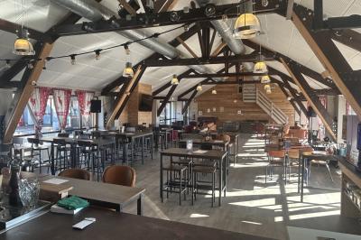 Salle de restaurant lumineuse avec poutres apparentes, grandes fenêtres, tables en bois et chaises hautes.
