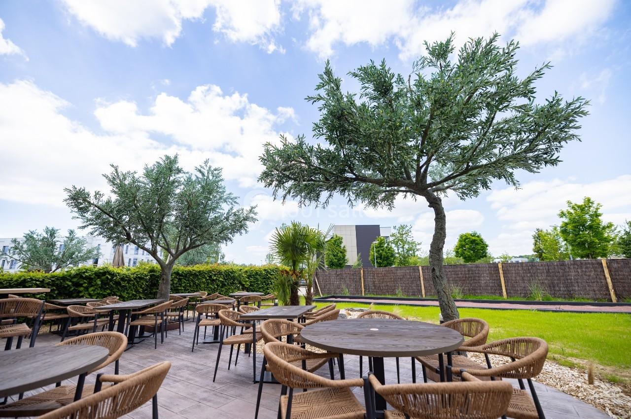 Terrasse extérieure avec tables et chaises en rotin, entourée de verdure et d'arbres sous un ciel partiellement nuageux.