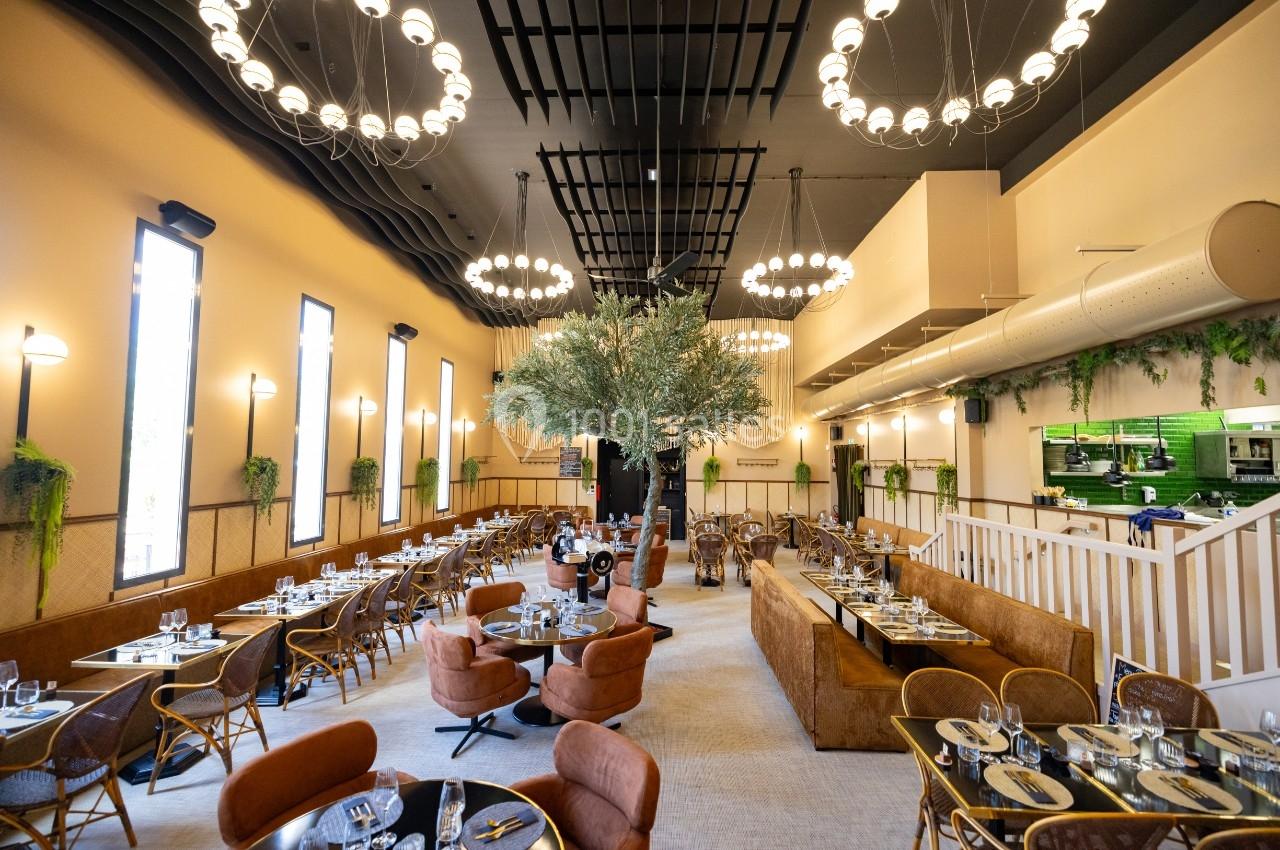 Salle de restaurant lumineuse avec tables dressées, chaises en rotin, arbre central et décoration végétale.