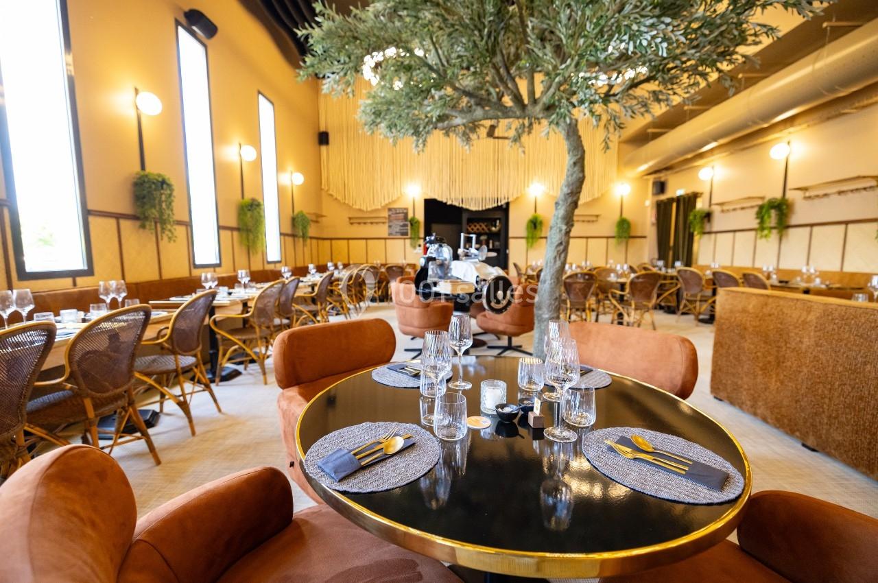 Salle de restaurant élégante avec tables dressées, chaises en velours, grand arbre central et lumière naturelle.