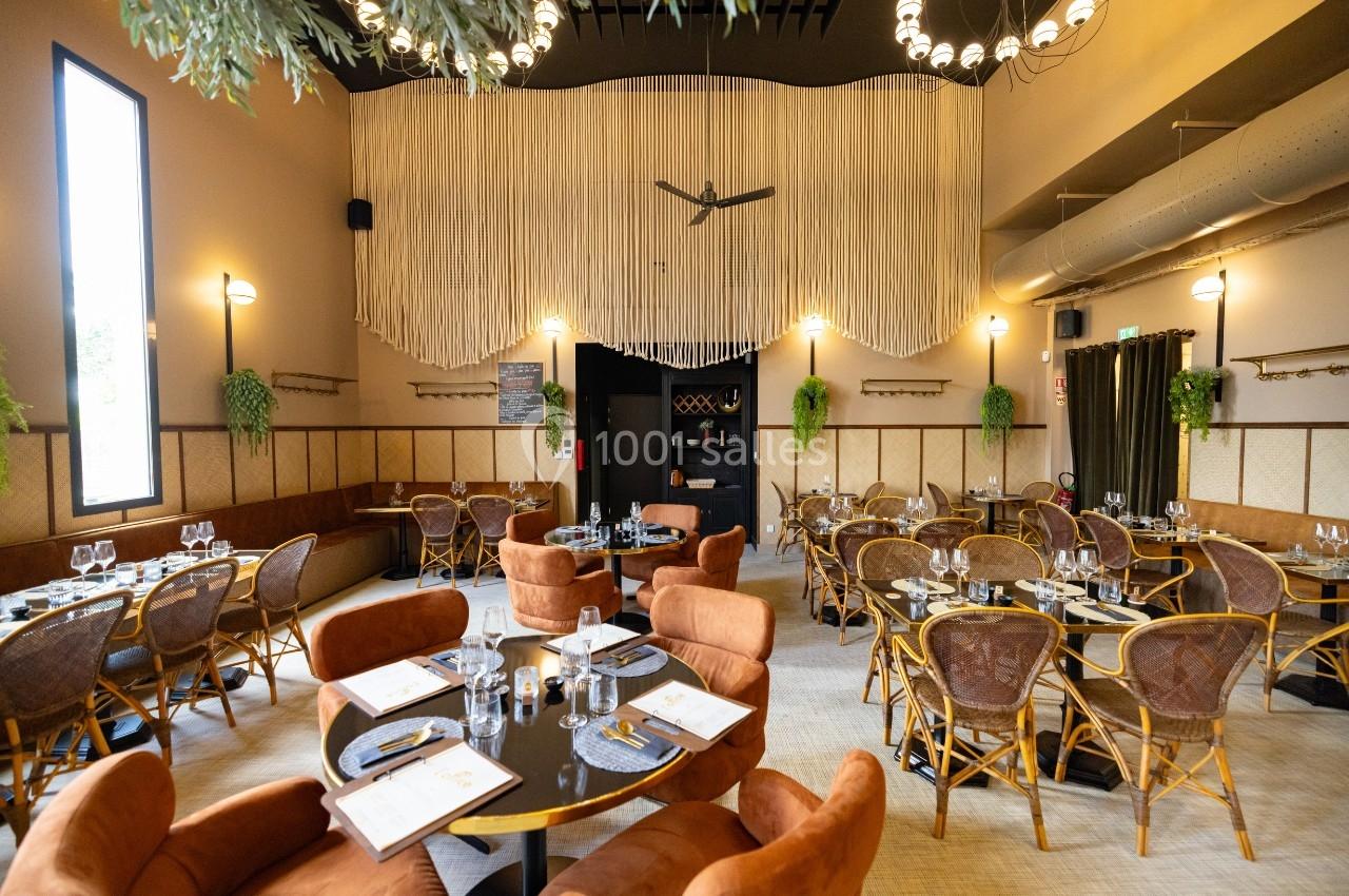 Salle de restaurant chaleureuse avec tables dressées, chaises en rotin et décoration végétale suspendue.