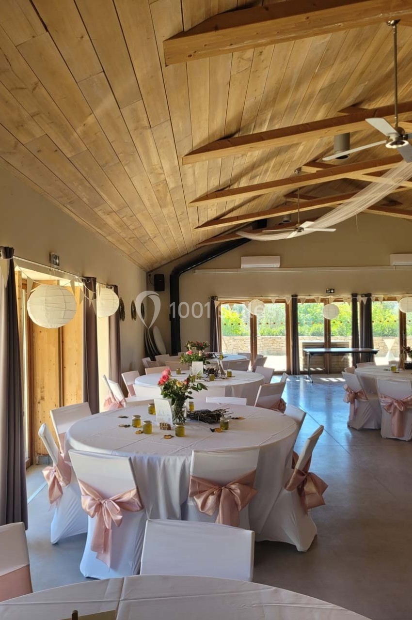 Salle lumineuse avec tables rondes décorées de nappes blanches et rubans roses, sous un plafond en bois.