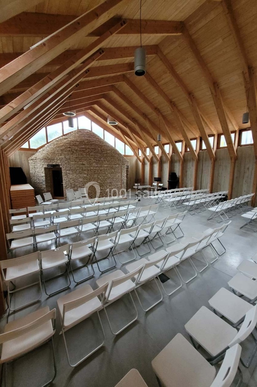 Salle lumineuse avec charpente en bois, murs en pierre et rangées de chaises blanches alignées.