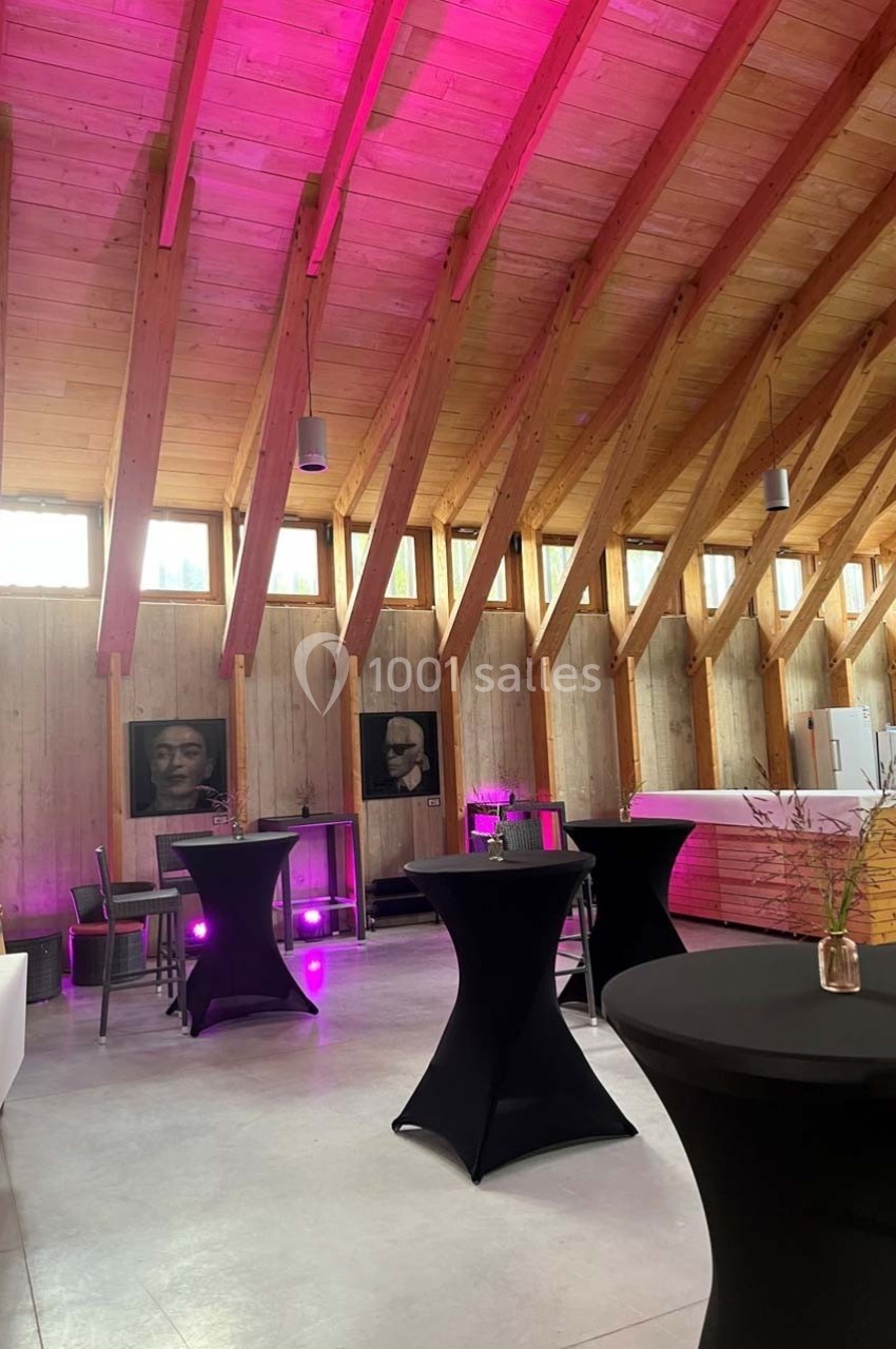 Salle en bois avec plafond voûté, tables hautes noires, éclairage rose et portraits accrochés aux murs.