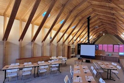 Salle de réunion lumineuse avec tables en U, chaises en bois, tonneau décoratif et étagères murales garnies de bouteilles.