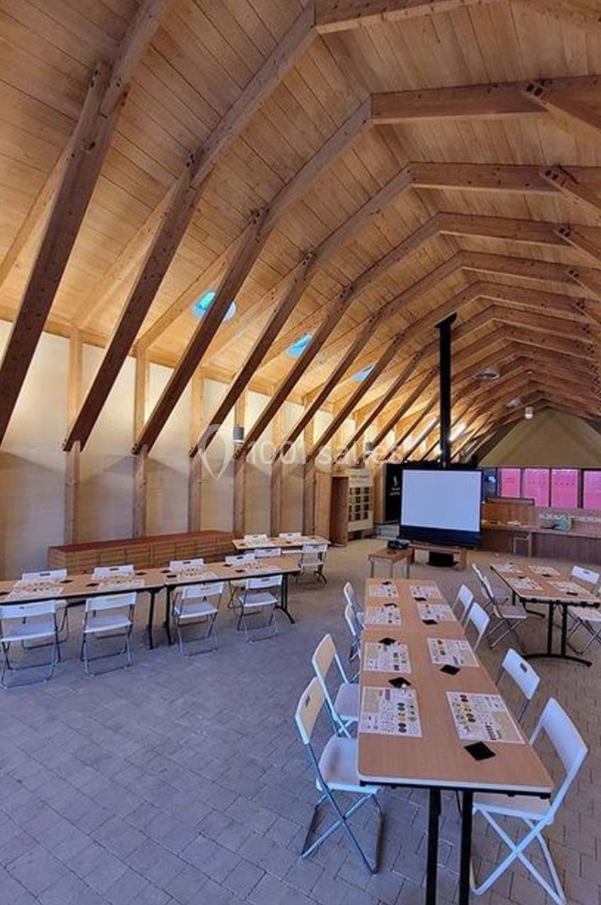 Salle en bois avec plafond voûté, tables alignées avec des documents et chaises pliantes, écran de projection au fond.