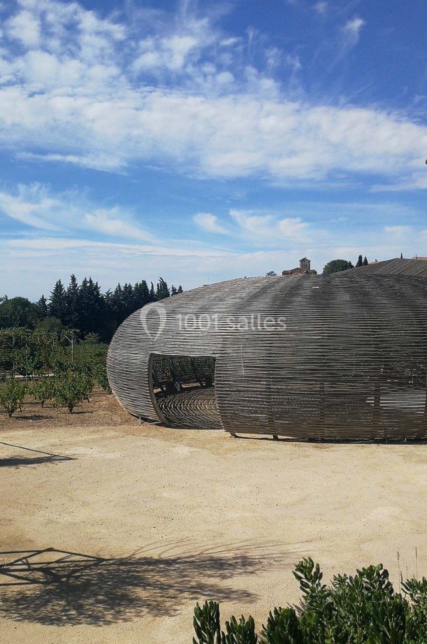 Structure architecturale en bois de forme ovale dans un paysage extérieur avec ciel bleu et végétation.