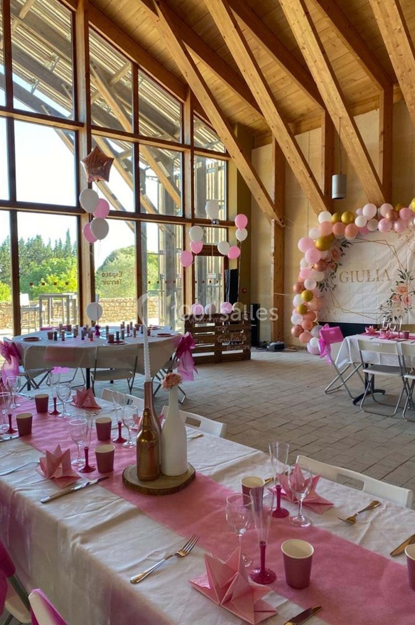 Salle décorée pour une fête avec tables dressées, ballons colorés et grande baie vitrée donnant sur un extérieur verdoyant.