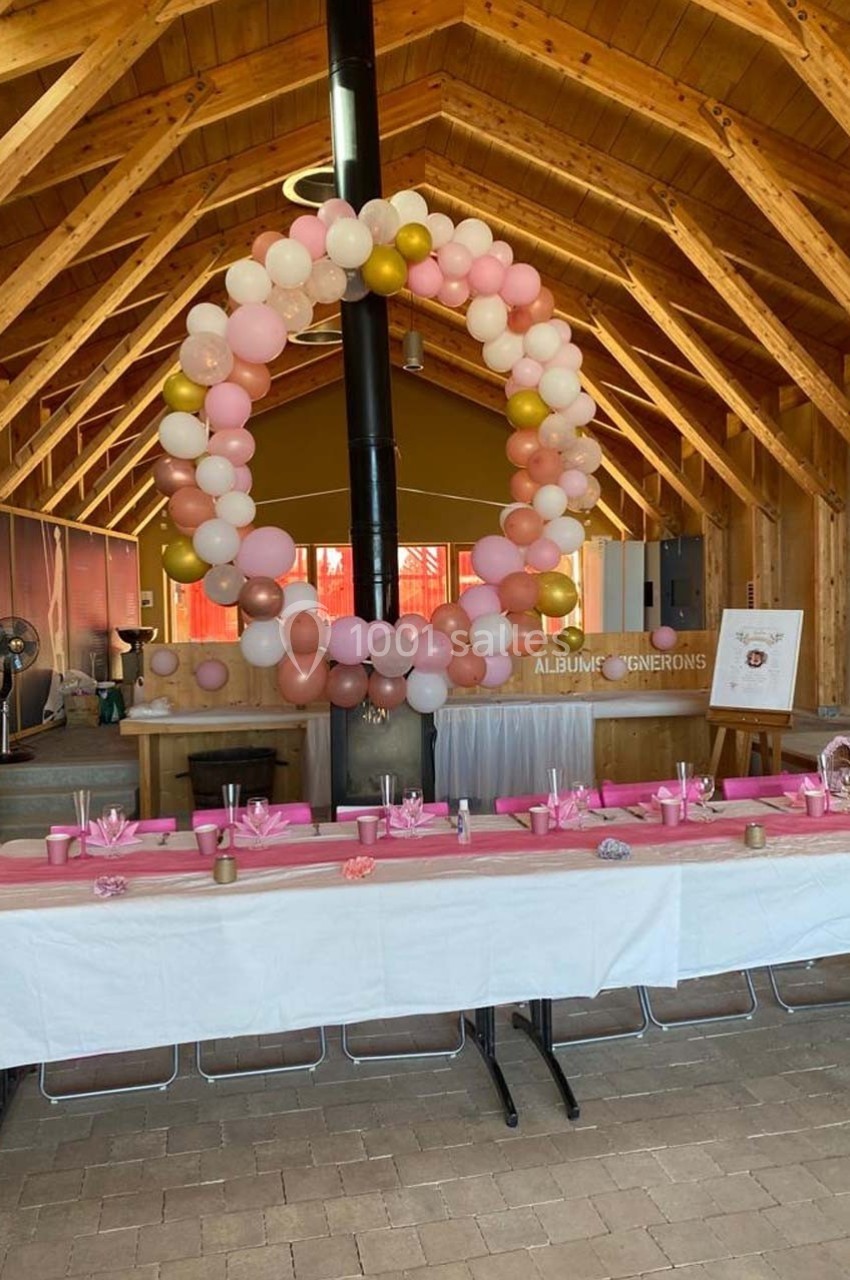 Décoration de salle en bois avec une table dressée et une arche de ballons roses, blancs et dorés au centre.