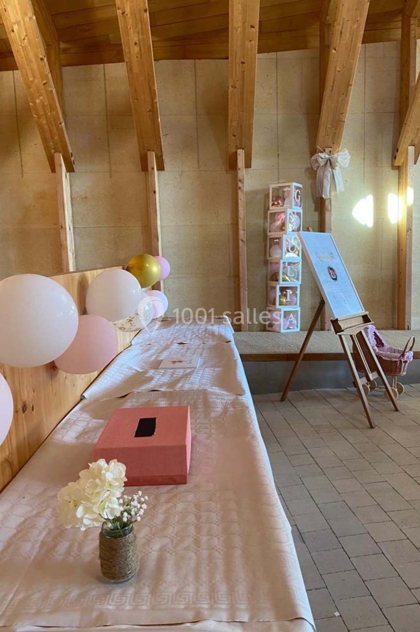 Salle décorée avec des ballons roses et blancs, une table avec nappes blanches, une urne rose et un chevalet avec photo.