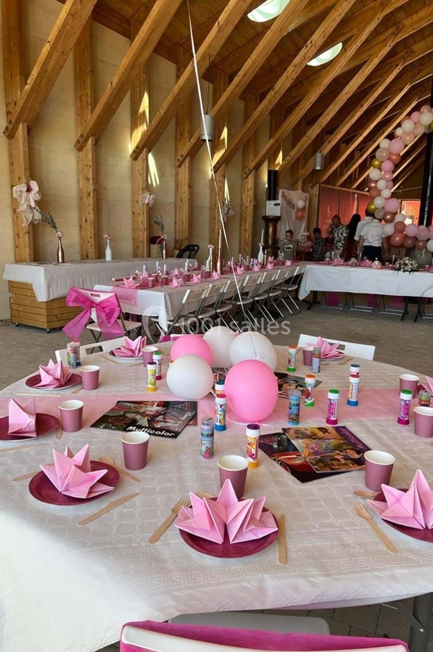Salle décorée pour une fête avec tables dressées, pliages roses, ballons et guirlande en arrière-plan.
