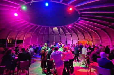 Salle de réunion lumineuse avec tables en U, chaises en bois, tonneau décoratif et étagères murales garnies de bouteilles.