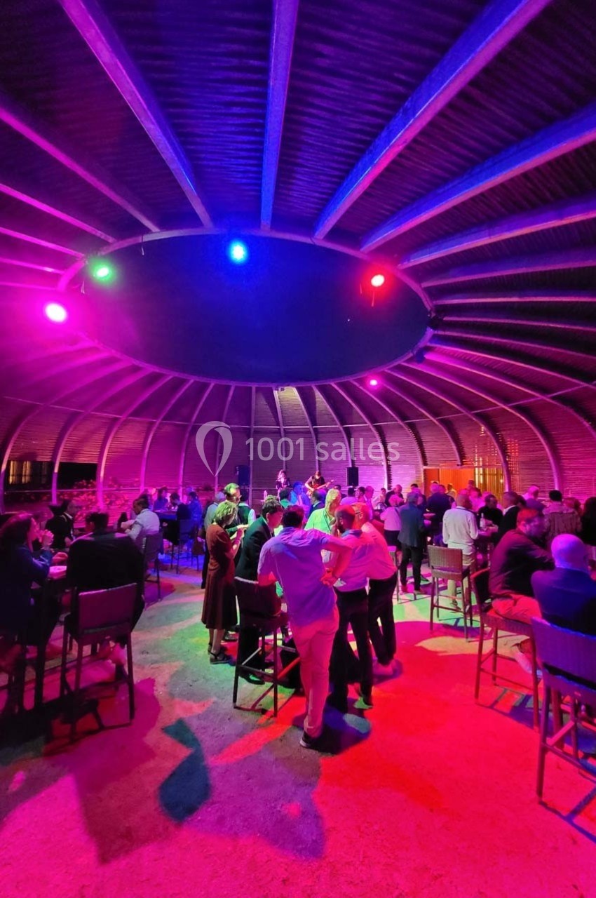 Des personnes rassemblées dans une salle circulaire éclairée par des lumières colorées, discutant et partageant un moment…