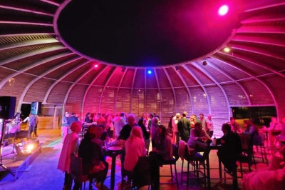 Salle de réunion lumineuse avec tables en U, chaises en bois, tonneau décoratif et étagères murales garnies de bouteilles.