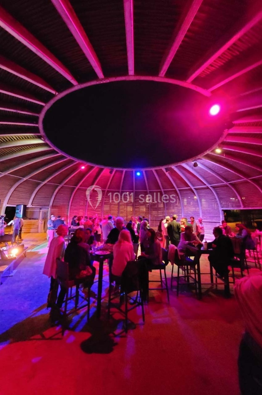Salle circulaire avec toit en bois, éclairée par des lumières colorées, occupée par des personnes debout et assises.