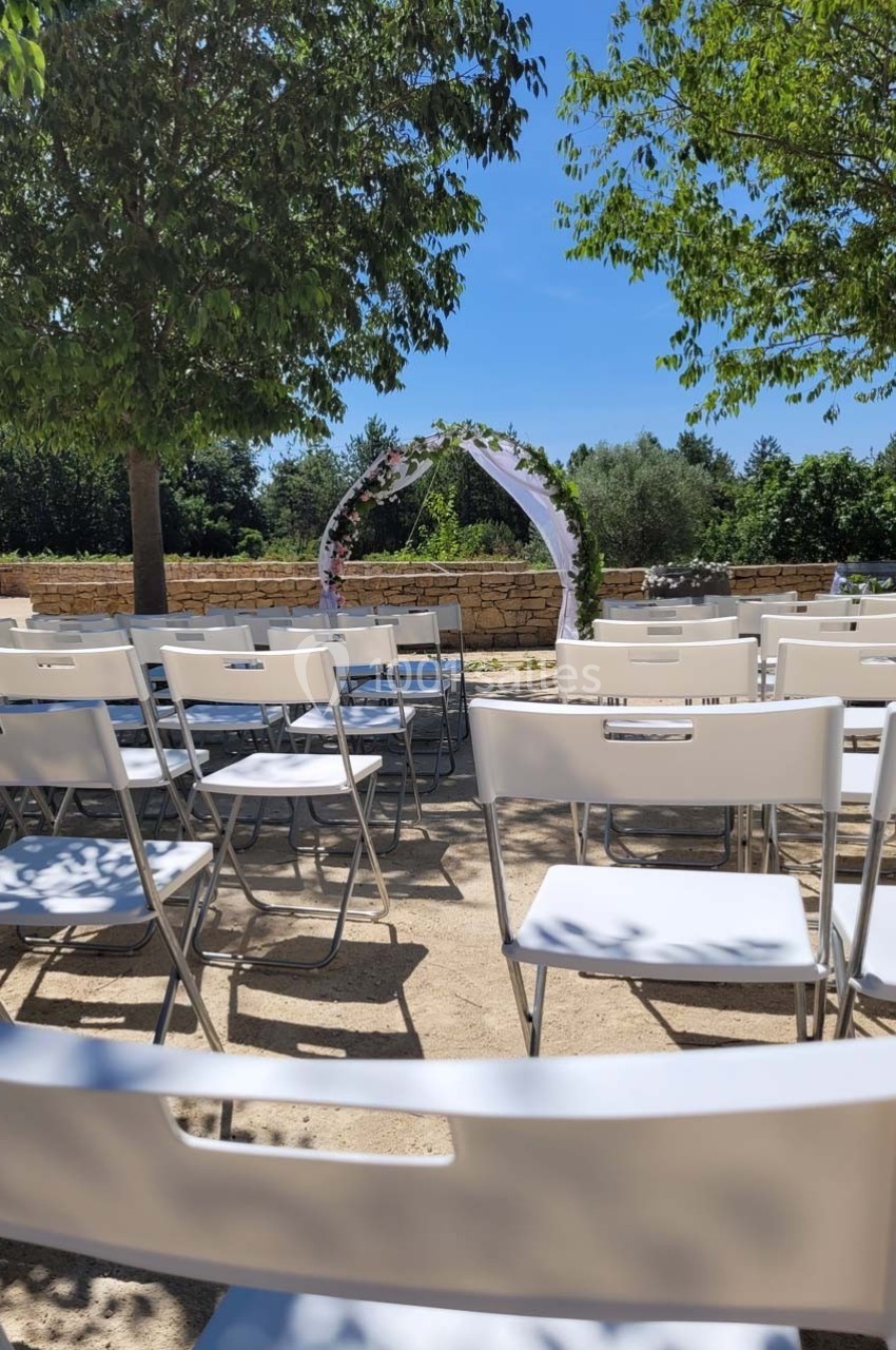 Chaises blanches disposées en rangées face à une arche décorée de fleurs dans un cadre extérieur ensoleillé.