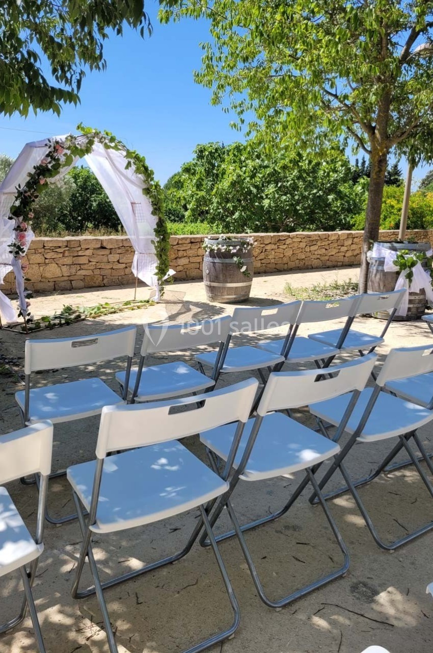 Chaises blanches alignées en extérieur devant une arche décorée de fleurs et de tissu, sous un ciel dégagé.