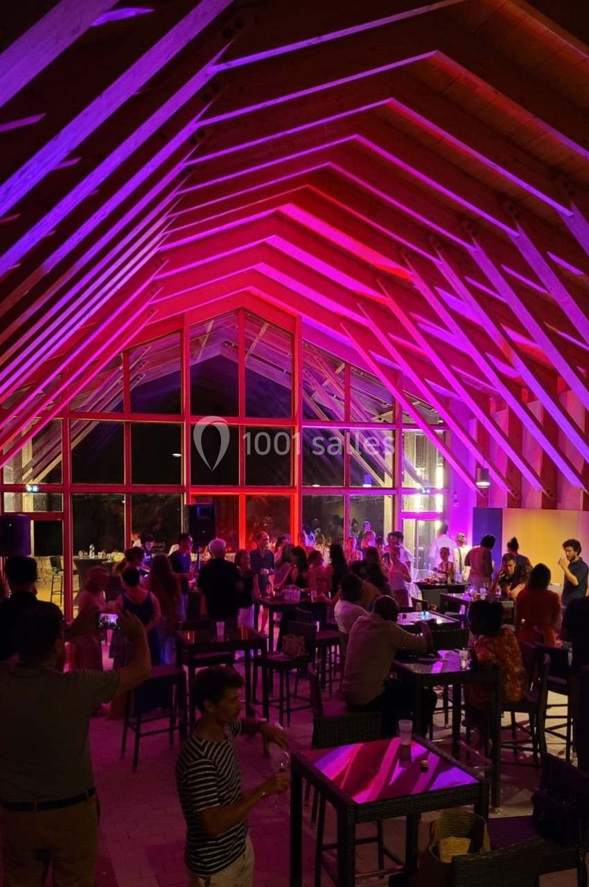 Salle en bois avec un plafond voûté, éclairée par des lumières roses, accueillant des personnes debout et assises.
