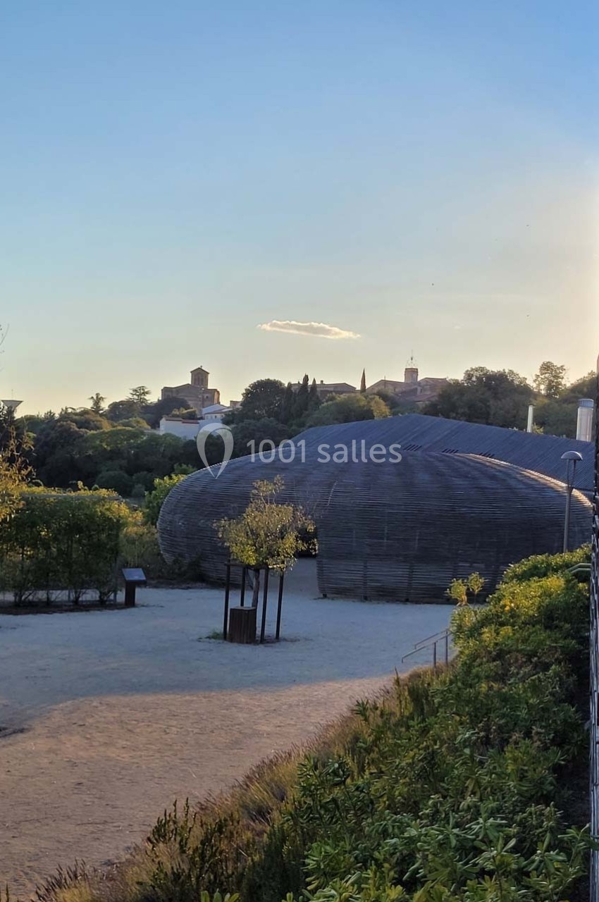 Vue d'un bâtiment en forme de dôme entouré de végétation, avec un village et un ciel dégagé en arrière-plan.