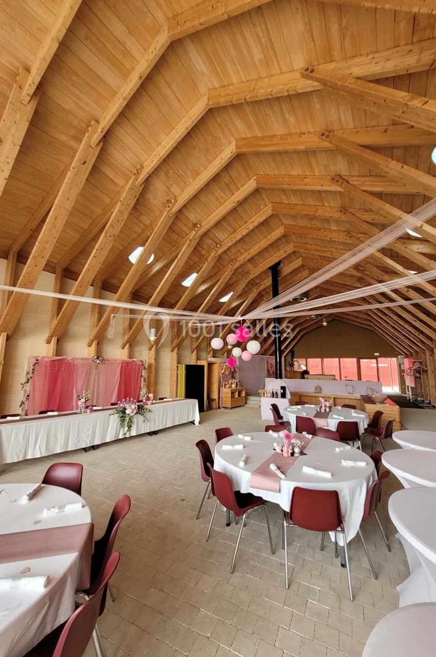Salle de réception en bois avec tables rondes décorées, guirlandes suspendues et espace buffet en arrière-plan.