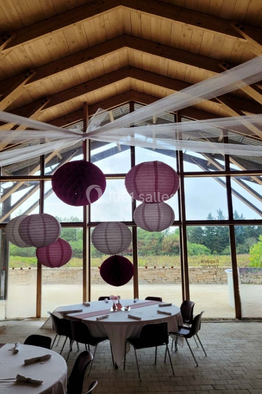 Salle avec charpente en bois, tables rondes dressées et lanternes suspendues devant de grandes baies vitrées.