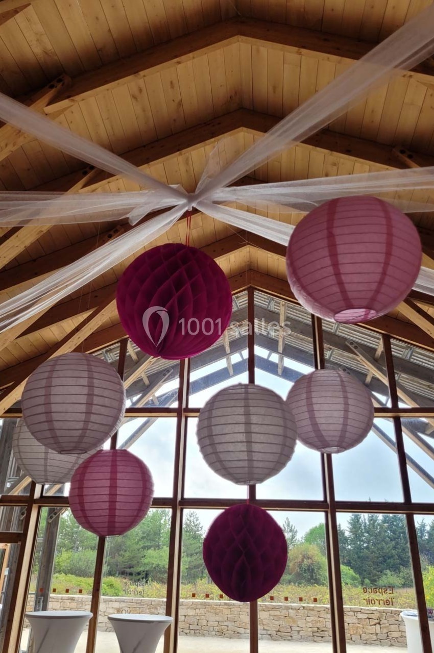 Lampions roses et blancs suspendus au plafond en bois d'une salle avec de grandes fenêtres donnant sur un jardin.