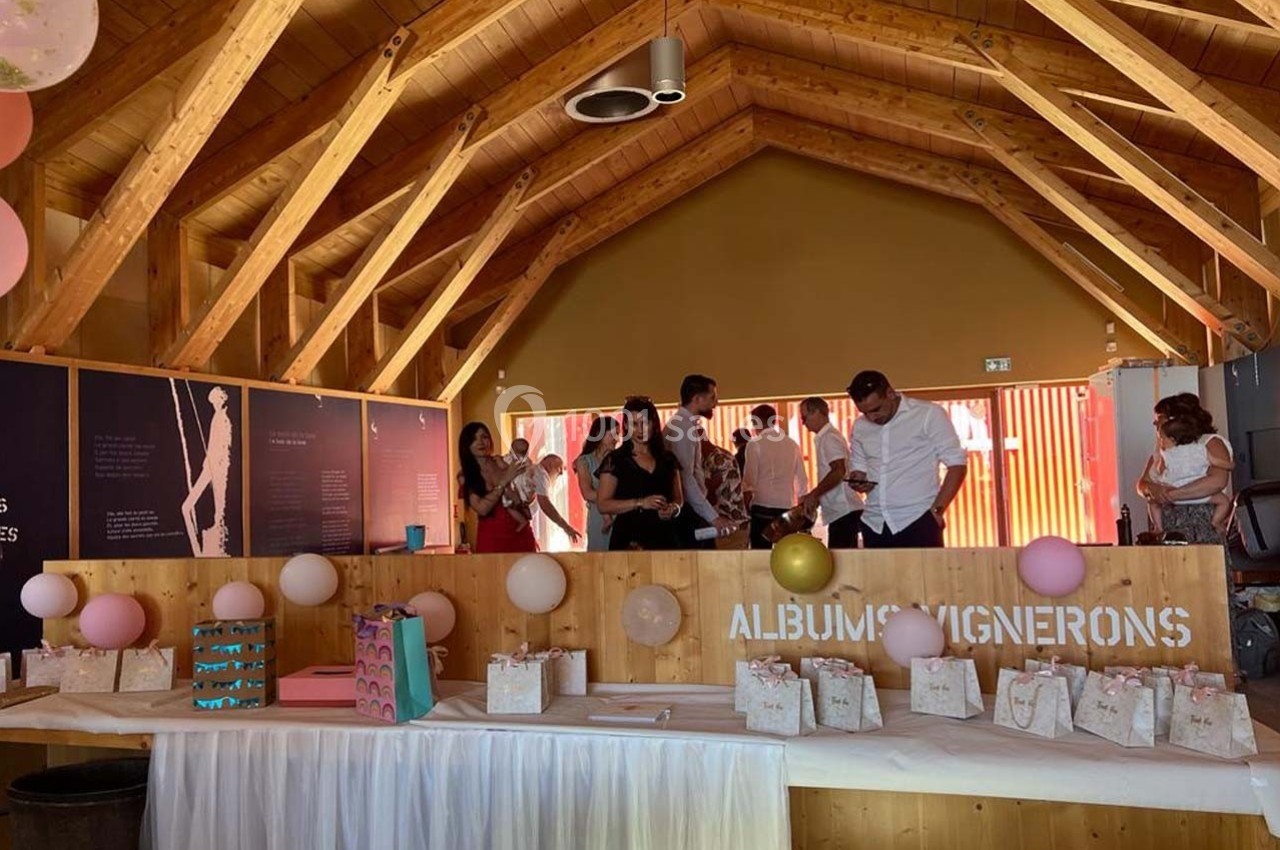 Salle en bois décorée pour un événement, avec des ballons, des cadeaux sur une table et des personnes discutant en arrière…