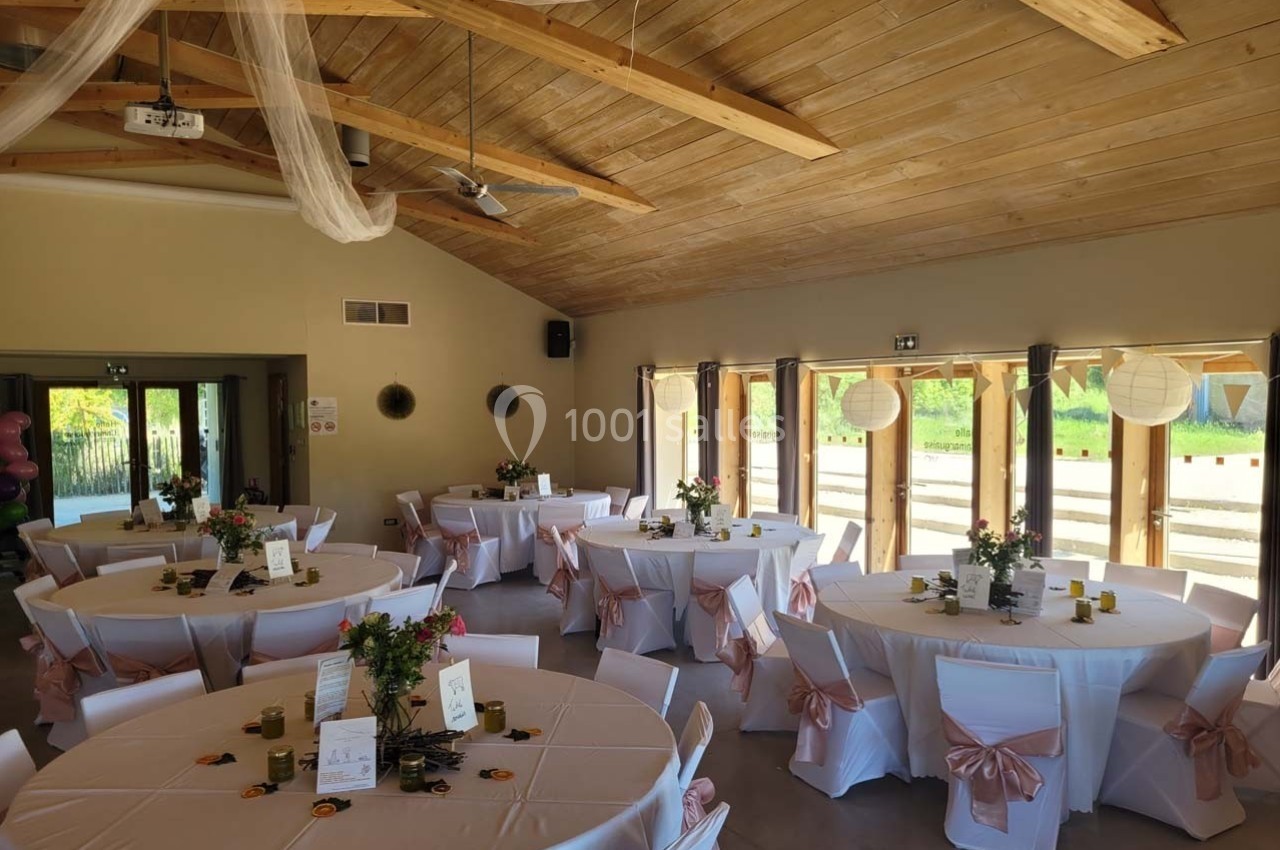Salle de réception décorée avec des tables rondes, nappes blanches, chaises ornées de rubans roses et arrangements floraux.