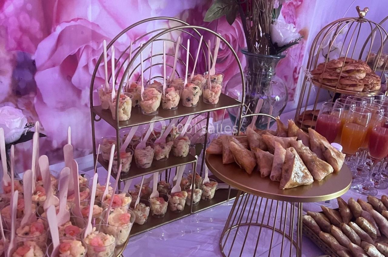 Buffet avec verrines, samoussas et jus de fruits, présenté sur des plateaux dorés devant un fond floral rose.