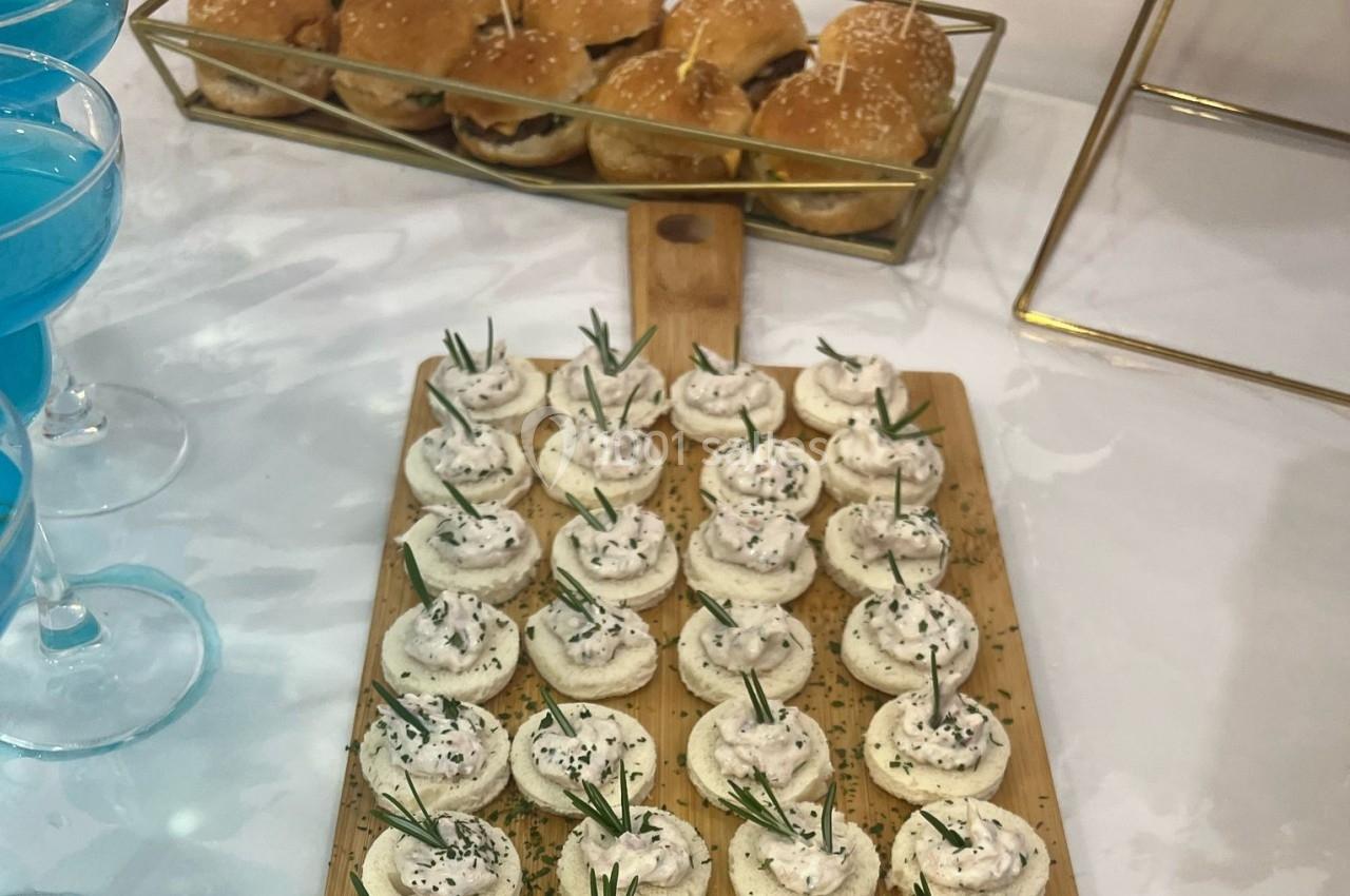 Plateau de canapés garnis de crème et herbes fraîches, avec des mini-burgers et des boissons bleues en arrière-plan.