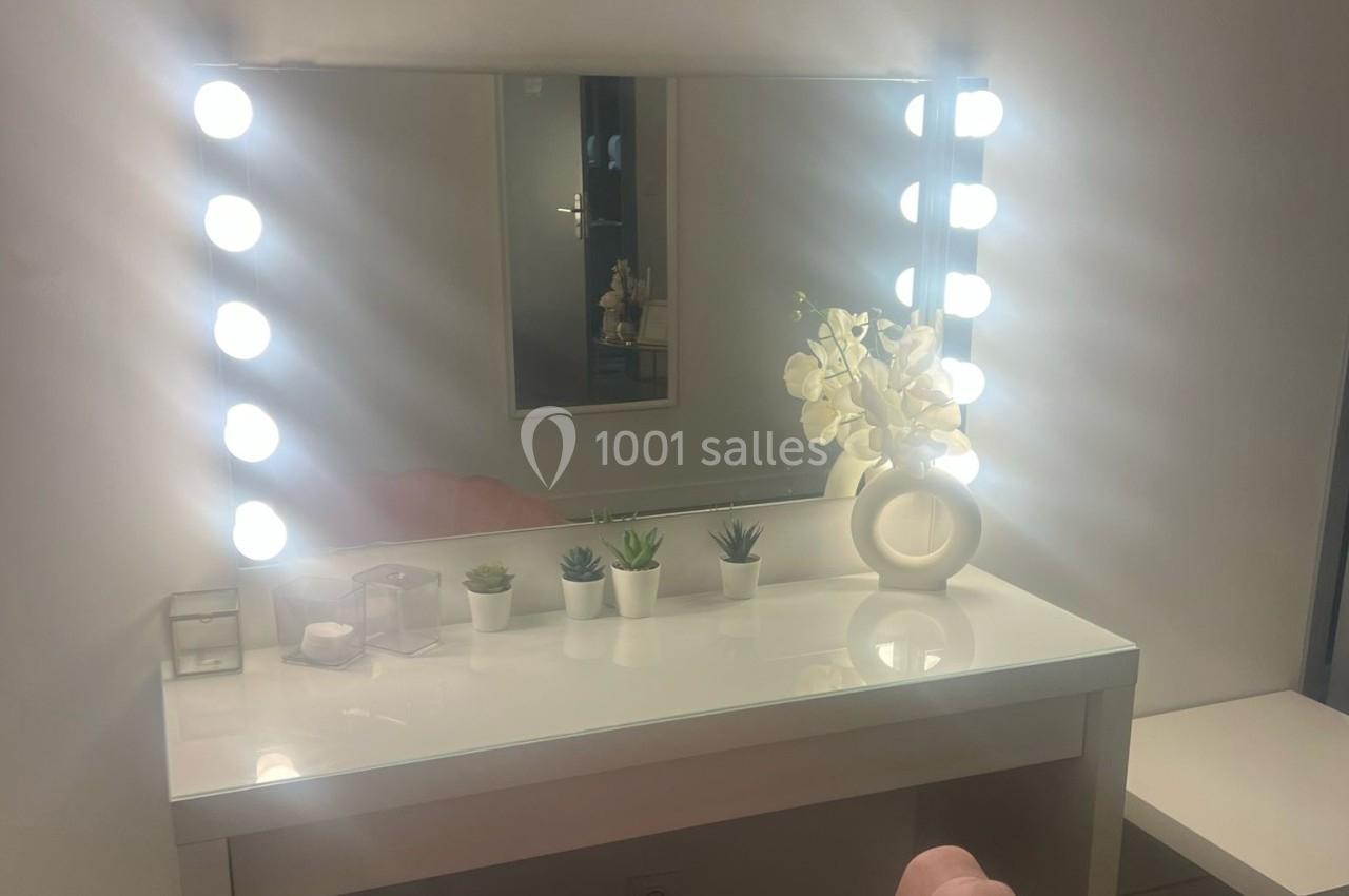 Miroir éclairé par des ampoules, posé sur une coiffeuse blanche avec des plantes et un vase décoratif.