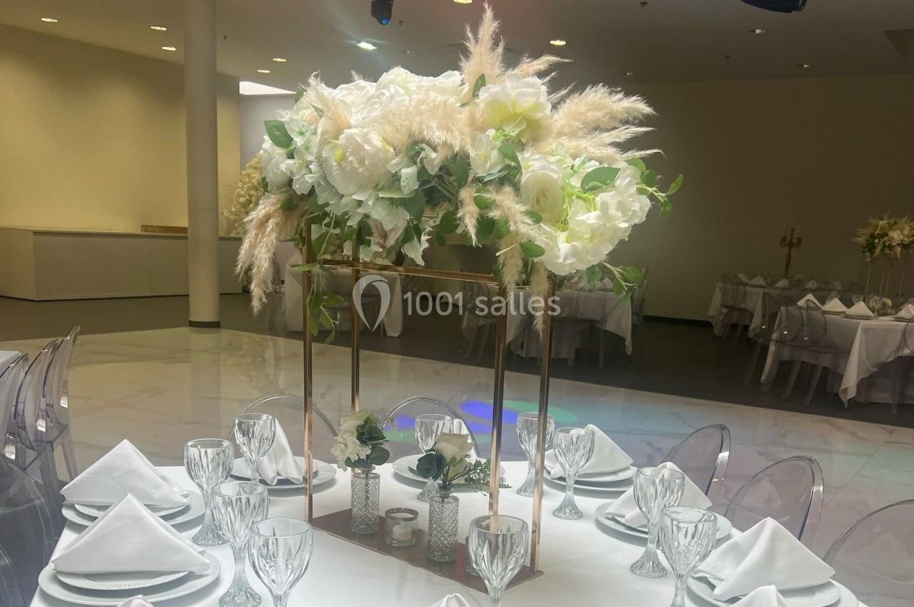 Centre de table élégant avec fleurs blanches, pampas et feuillage, entouré de verres et serviettes pliées sur une table…