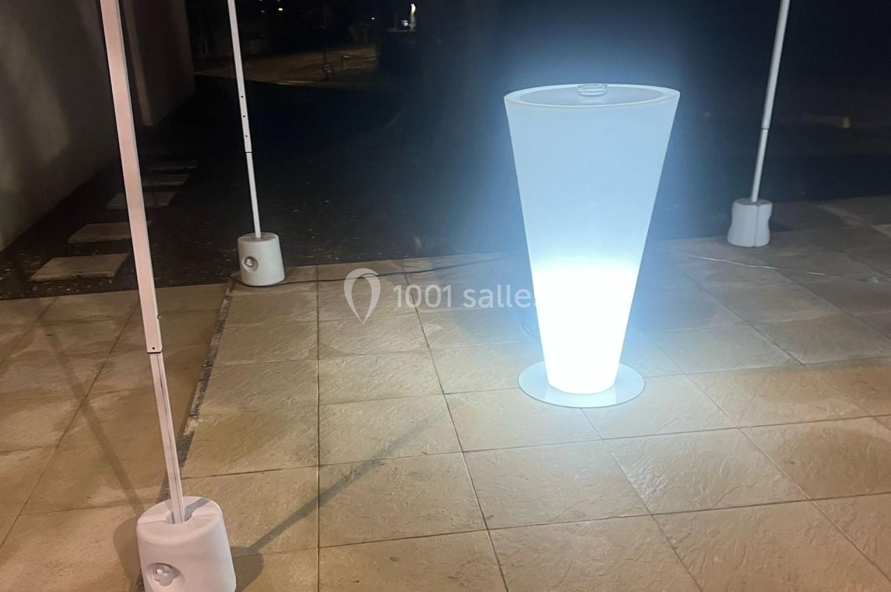 Lampe conique lumineuse blanche éclairant une terrasse en dalles, entourée de poteaux avec socles.