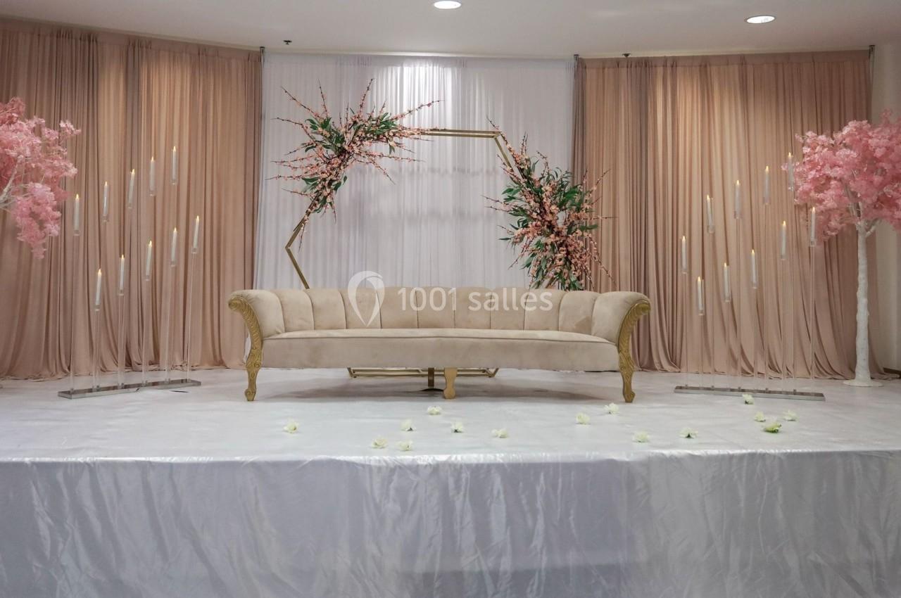 Canapé beige avec accoudoirs dorés, décoré de fleurs roses et blanches, devant des rideaux beiges et blancs.