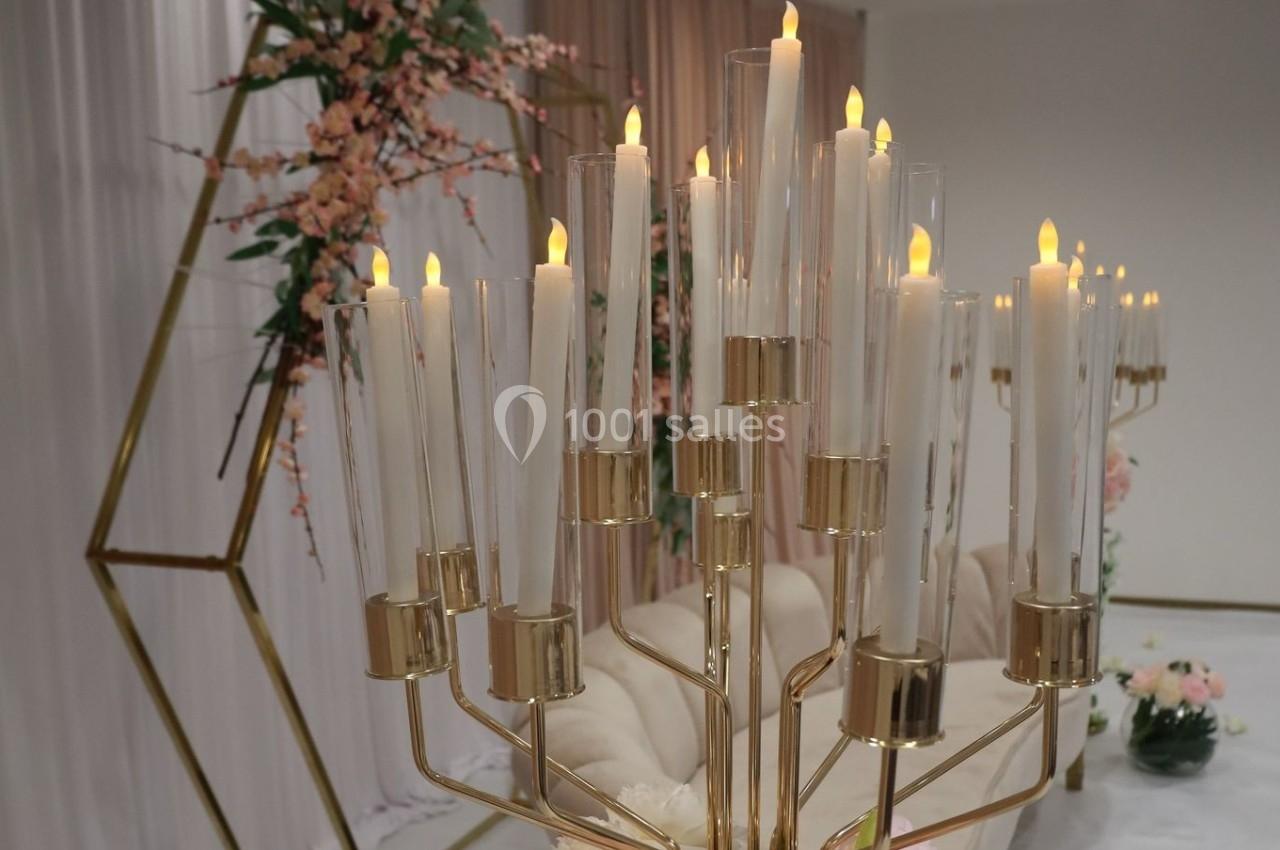 Chandelier doré avec bougies allumées, devant une décoration florale et un fond de rideaux roses et blancs.