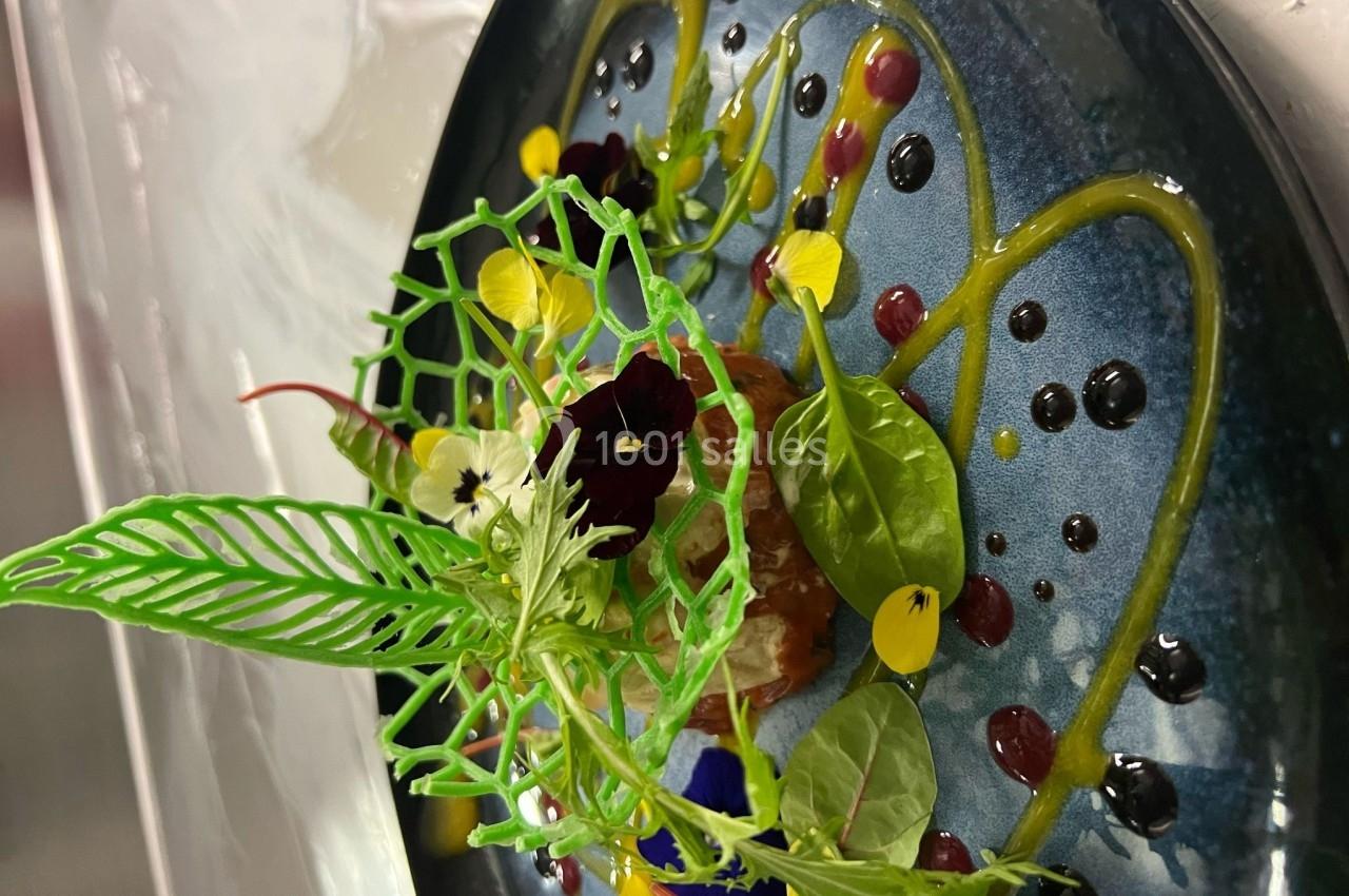 Assiette gastronomique décorée de fleurs comestibles, feuilles et sauces colorées sur un fond bleu.